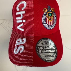 May include: A red snapback cap featuring the word Chivas embroidered in white on the left side. The right side displays the Club Deportivo Guadalajara crest. A circular sticker on the brim reads High Quality Premium Snapback Classics.