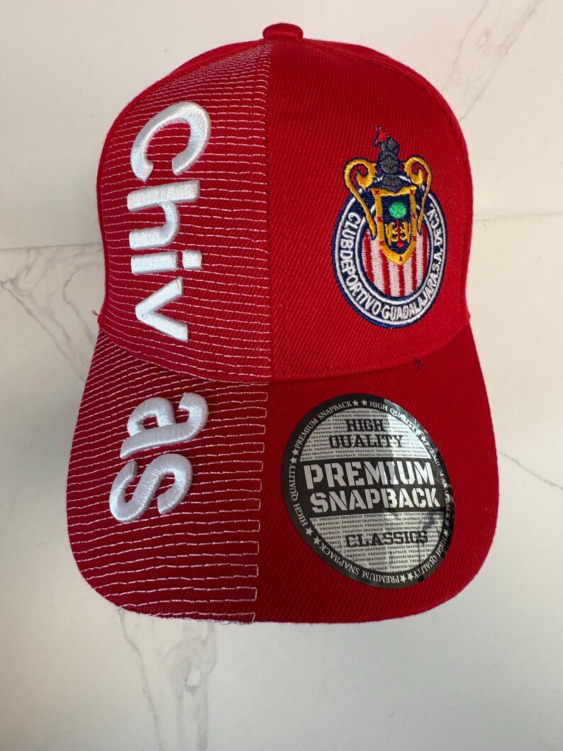 May include: A red snapback cap featuring the word Chivas embroidered in white on the left side. The right side displays the Club Deportivo Guadalajara crest. A circular sticker on the brim reads High Quality Premium Snapback Classics.