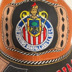 May include: An orange baseball cap featuring a central circular patch with a black leather-like background. The patch displays a colourful embroidered crest with a helmet, shield, and the text Club Deportivo Guadalajara. Tan leather accents frame the patch.