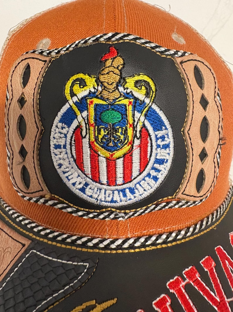 May include: An orange baseball cap featuring a central circular patch with a black leather-like background. The patch displays a colourful embroidered crest with a helmet, shield, and the text Club Deportivo Guadalajara. Tan leather accents frame the patch.