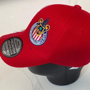 May include: A bright red baseball cap featuring the embroidered Club Deportivo Guadalajara logo on the side. The cap includes a circular black sticker on the brim with the text Premium Snapback and High Quality Classics printed in white.