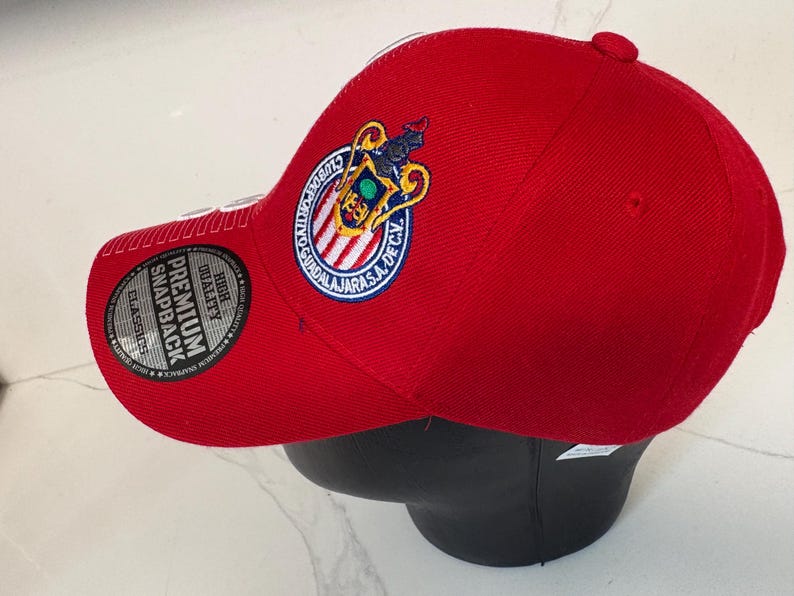 May include: A bright red baseball cap featuring the embroidered Club Deportivo Guadalajara logo on the side. The cap includes a circular black sticker on the brim with the text Premium Snapback and High Quality Classics printed in white.