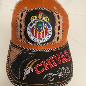 May include: An orange and tan trucker hat featuring a central patch with the Chivas Guadalajara crest. The black brim displays the word CHIVAS in red embroidery, a white goat head graphic, and decorative tan leather-like accents with stitching.