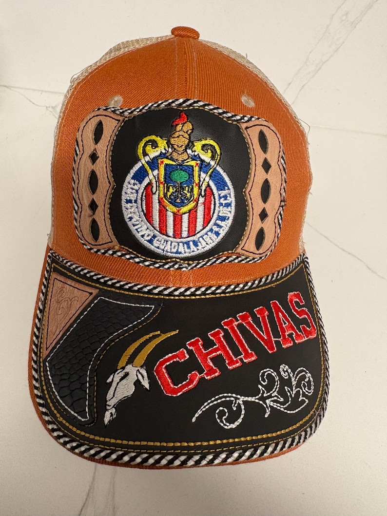 May include: An orange and tan trucker hat featuring a central patch with the Chivas Guadalajara crest. The black brim displays the word CHIVAS in red embroidery, a white goat head graphic, and decorative tan leather-like accents with stitching.