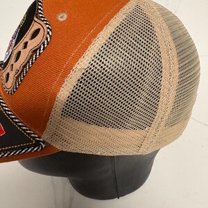 May include: A trucker cap featuring a burnt orange front panel and a beige mesh back. The front displays a black patch with a decorative braided trim and a tan leather-like accent piece with cutouts. The cap has a matching orange button on top.