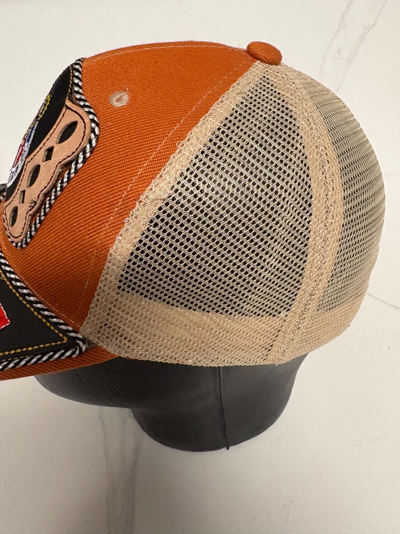 May include: A trucker cap featuring a burnt orange front panel and a beige mesh back. The front displays a black patch with a decorative braided trim and a tan leather-like accent piece with cutouts. The cap has a matching orange button on top.