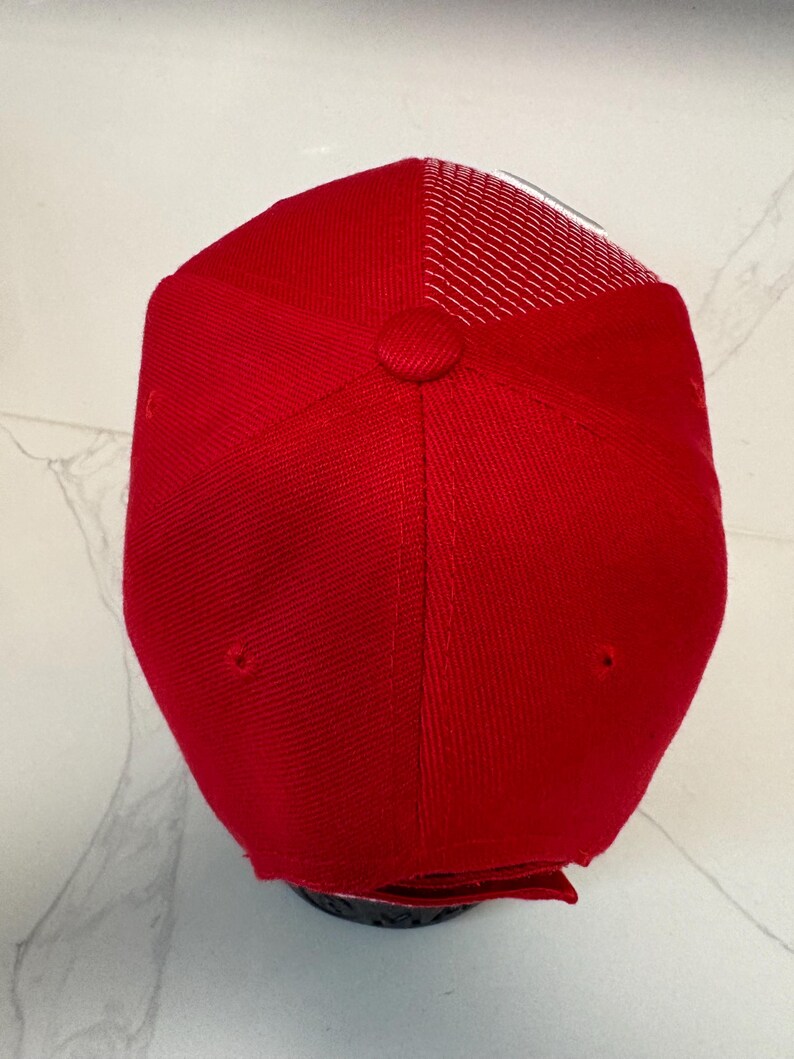 May include: A bright red baseball cap featuring a structured front panel with a fabric-covered button at the crown and a breathable white mesh back panel. The cap has a classic six-panel construction with stitched eyelets for ventilation.