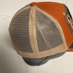May include: A trucker cap featuring a solid orange front panel and a beige mesh back panel. The cap includes a small orange button at the top centre where the panels meet. A partial view of a dark patch with decorative stitching is visible on the side.