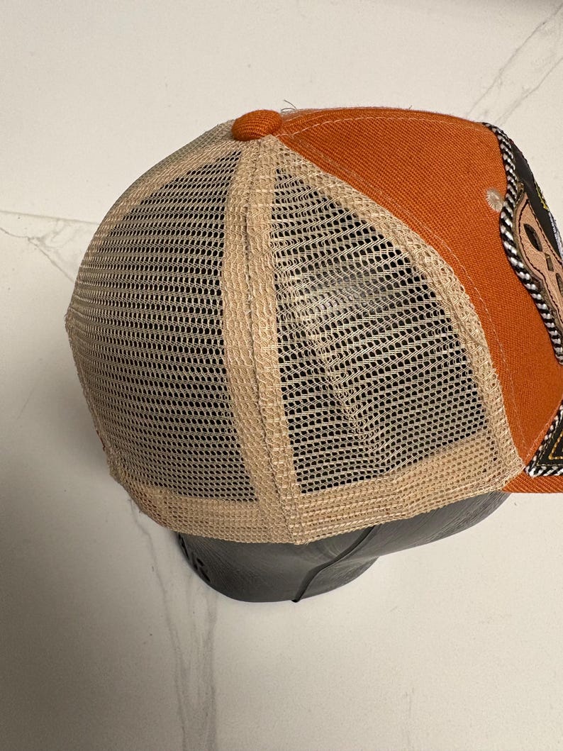 May include: A trucker cap featuring a solid orange front panel and a beige mesh back panel. The cap includes a small orange button at the top centre where the panels meet. A partial view of a dark patch with decorative stitching is visible on the side.