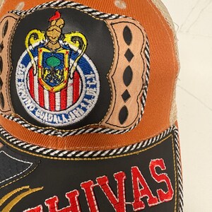 May include: An orange and tan trucker cap featuring a circular patch with the Chivas Guadalajara crest, surrounded by tan leather-like accents. The black brim displays the word CHIVAS in bold red embroidered letters with white stitching.