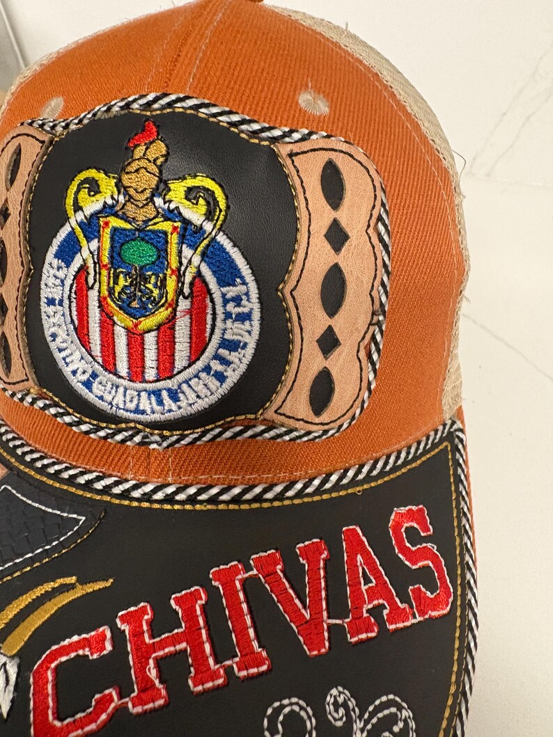 May include: An orange and tan trucker cap featuring a circular patch with the Chivas Guadalajara crest, surrounded by tan leather-like accents. The black brim displays the word CHIVAS in bold red embroidered letters with white stitching.
