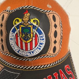 May include: An orange baseball cap featuring a central circular patch with a crest displaying a tree, red and white stripes, and a gold helmet. The patch is framed by tan leather-like accents and the word CHIVAS is embroidered in red on the black brim.