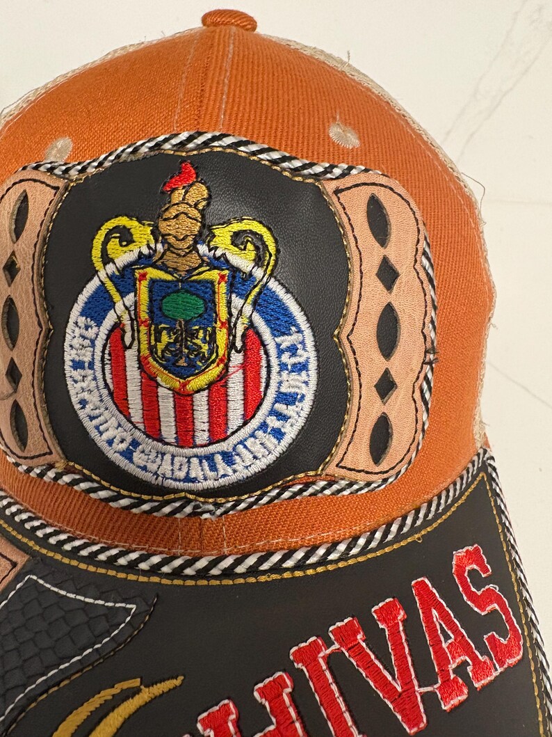 May include: An orange baseball cap featuring a central circular patch with a crest displaying a tree, red and white stripes, and a gold helmet. The patch is framed by tan leather-like accents and the word CHIVAS is embroidered in red on the black brim.