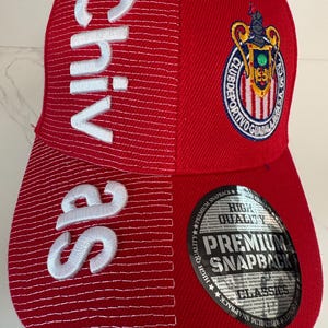 May include: A red snapback cap featuring white embroidered text that reads Chivas vertically on the left side. The cap displays the Club Deportivo Guadalajara crest on the upper right and includes a circular black sticker on the brim with the text High Quality Premium Snapback Classics.