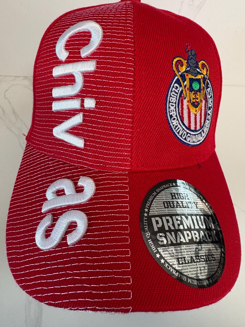 May include: A red snapback cap featuring white embroidered text that reads Chivas vertically on the left side. The cap displays the Club Deportivo Guadalajara crest on the upper right and includes a circular black sticker on the brim with the text High Quality Premium Snapback Classics.