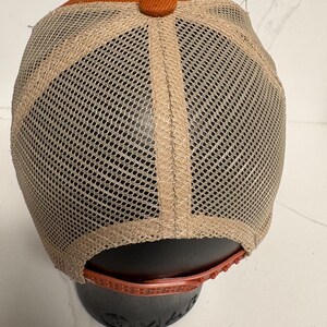 May include: A baseball cap featuring a solid burnt orange fabric crown and a matching fabric button at the top. The back and side panels are constructed from a tan mesh material. An adjustable plastic snapback closure in burnt orange is at the base.