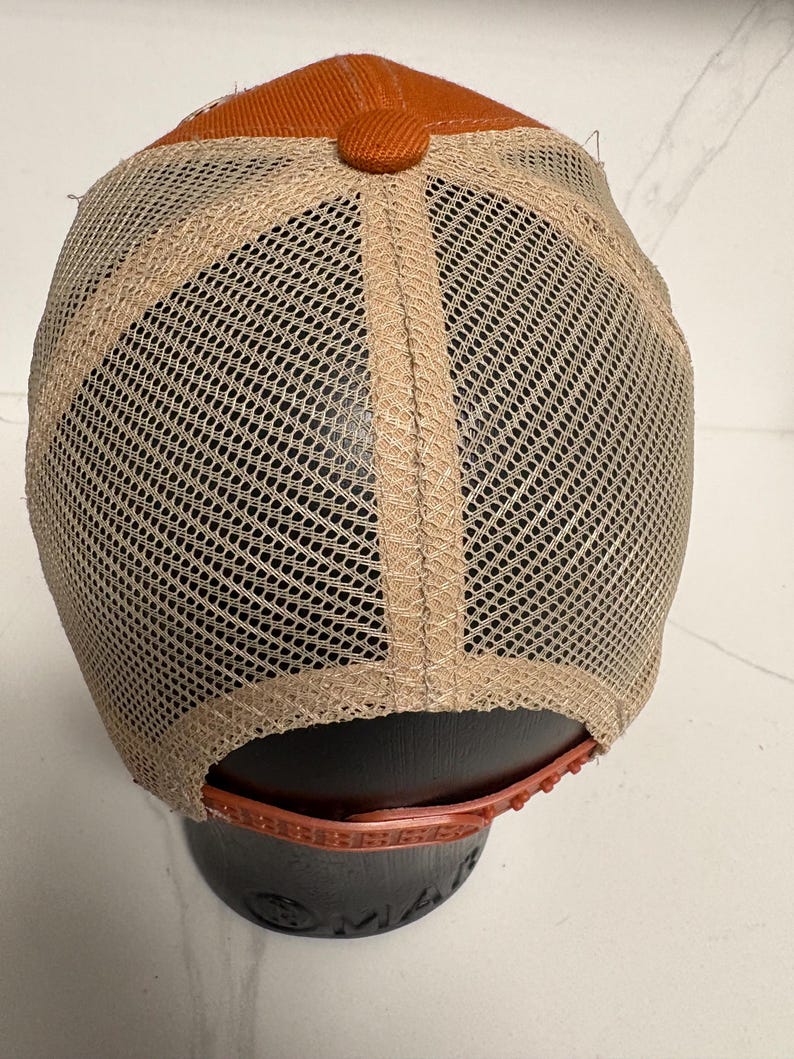 May include: A baseball cap featuring a solid burnt orange fabric crown and a matching fabric button at the top. The back and side panels are constructed from a tan mesh material. An adjustable plastic snapback closure in burnt orange is at the base.