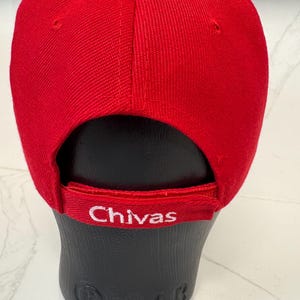 May include: A bright red baseball cap featuring a structured crown and a curved brim. The back strap is adjustable and displays the word Chivas embroidered in white thread. The cap is displayed on a black mannequin head form.