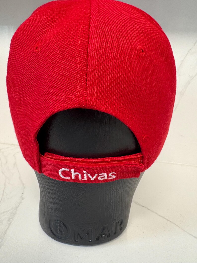 May include: A bright red baseball cap featuring a structured crown and a curved brim. The back strap is adjustable and displays the word Chivas embroidered in white thread. The cap is displayed on a black mannequin head form.