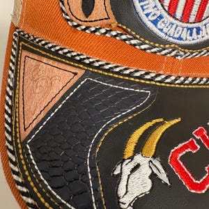 May include: An orange and black baseball cap featuring a circular embroidered patch with red and white stripes, a goat head emblem, and a triangular tan leather accent. The cap includes black and white braided trim and textured black panels.