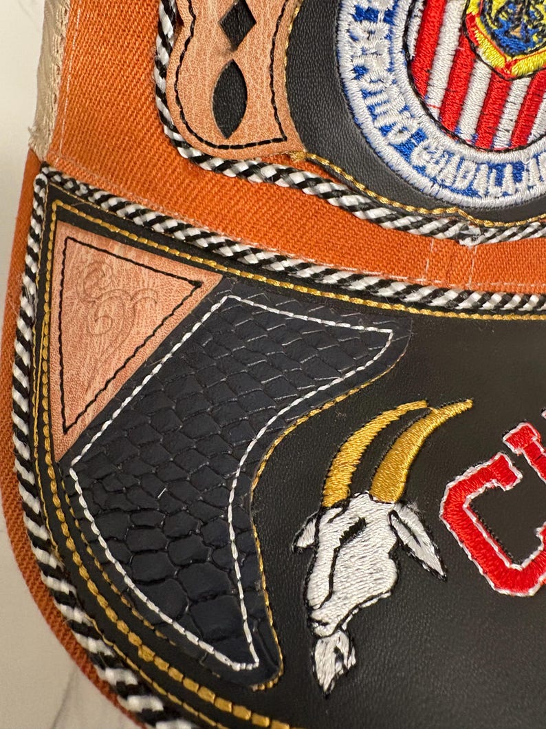 May include: An orange and black baseball cap featuring a circular embroidered patch with red and white stripes, a goat head emblem, and a triangular tan leather accent. The cap includes black and white braided trim and textured black panels.