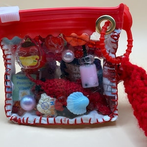 May include: A small, clear, rectangular pouch with red plastic trim and a red zipper closure. The pouch is filled with various charms, including a small sign that reads "Sending Positive Vibes," a pink boba tea charm, and a blue seashell.