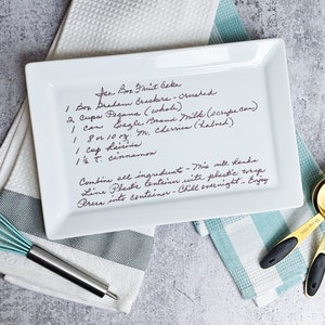 May include: A white rectangular ceramic plate with handwritten instructions for making a fruit cake. The instructions include ingredients such as graham crackers, pecans, evaporated milk, cherries, raisins, and cinnamon. The plate is sitting on a white and blue striped kitchen towel.