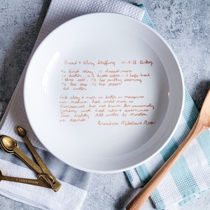 May include: A white ceramic plate with a handwritten recipe for bread and celery stuffing for a 14-18 lb turkey. The recipe includes ingredients like butter, bread cubes, celery, salt, sage, pepper, and hot water. The recipe is written in cursive and ends with "Grandma Mikolena's Mom".