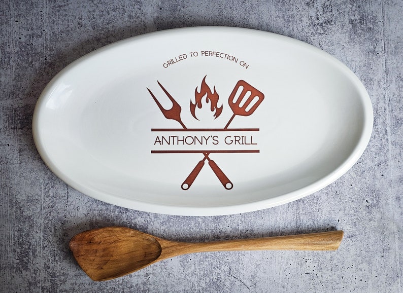 May include: White oval serving platter with a brown design of crossed grilling tools, a flame, and the text "Grilled to perfection on Anthony's Grill".
