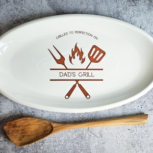 May include: White oval ceramic serving platter with a brown design of crossed grilling tools, a flame, and the text "Grilled to perfection on Dad's Grill".