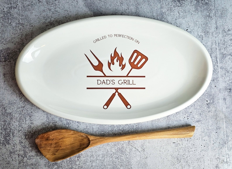 May include: White oval ceramic serving platter with a brown design of crossed grilling tools, a flame, and the text "Grilled to perfection on Dad's Grill".