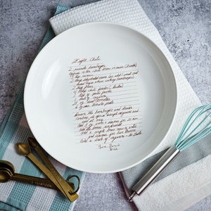 May include: A white ceramic serving bowl with a handwritten recipe for "Eagle Chile" printed on the inside. The recipe includes ingredients like hamburger, chilli powder, and tomato paste. The bowl is sitting on a blue and white striped tea towel with a set of measuring spoons and a whisk.