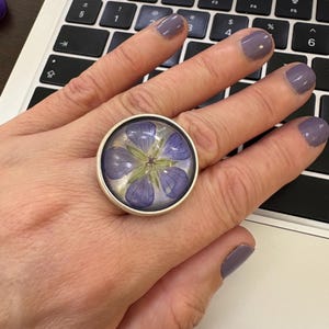 May include: A silver-toned ring with a large, round face. The ring's face features a preserved purple flower encased in clear resin. The flower's petals are visible, creating a unique, botanical jewelry piece.