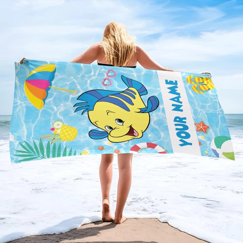 Puede incluir: Una toalla de playa rectangular con fondo azul de patr&oacute;n de agua, que presenta un gran pez de dibujos animados amarillo y azul, una sombrilla colorida, una bebida de pi&ntilde;a, chanclas, una estrella de mar y el texto YOUR NAME en letras azules.