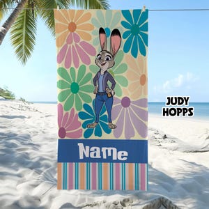 Może przedstawiać: Ręcznik plażowy z kresk&oacute;wkowym wizerunkiem Judy Hopps z Zootopii, otoczony dużymi, kolorowymi kwiatami. Ręcznik ma niebieski pasek na dole ze słowem "Name" w kolorze białym. Widoczny jest r&oacute;wnież tekst "JUDY HOPPS".