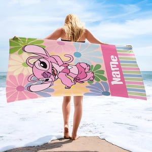 May include: A pink beach towel featuring a cartoon character with a floral background. The towel has a pink stripe with the word "Name" in white. The character is pink and the flowers are pink, yellow, green, and blue.