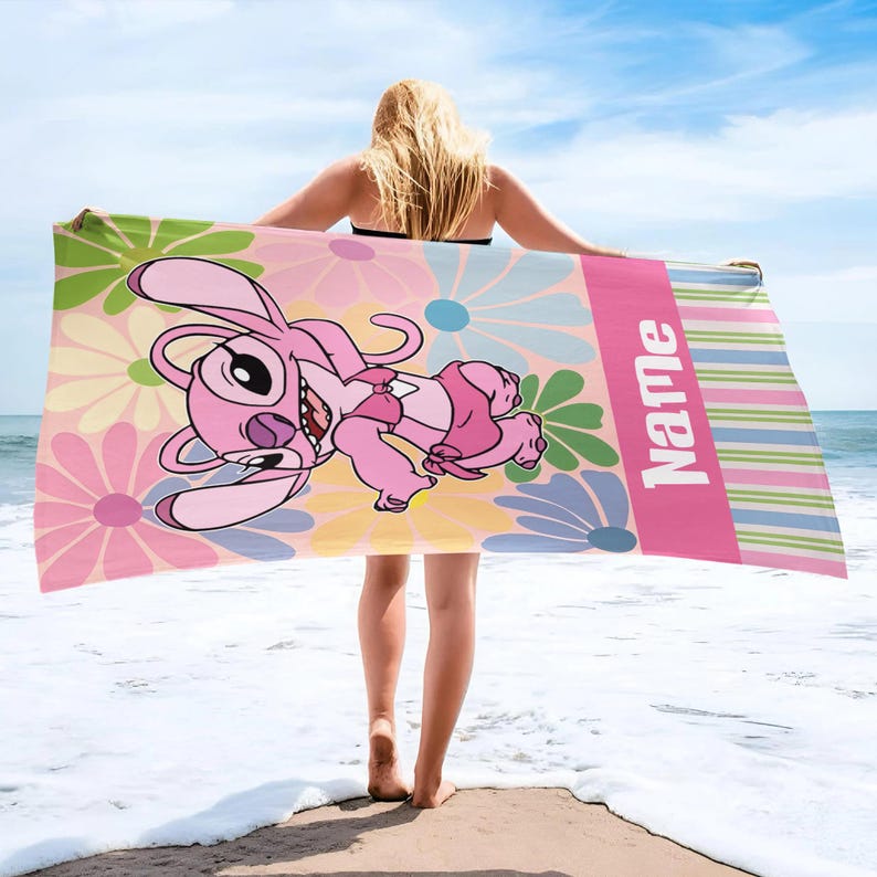 May include: A pink beach towel featuring a cartoon character with a floral background. The towel has a pink stripe with the word "Name" in white. The character is pink and the flowers are pink, yellow, green, and blue.