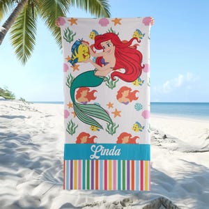 K&ouml;nnte beinhalten: Wei&szlig;es Strandtuch mit einer Meerjungfrau mit roten Haaren und einem gr&uuml;nen Schwanz, die einen gelben Fisch h&auml;lt. Das Handtuch hat einen blauen Streifen mit dem Namen "Linda" und einen bunten gestreiften Rand. Maritimes Design mit Seesternen und Muscheln.