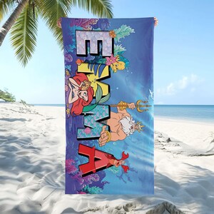 K&ouml;nnte beinhalten: Ein rechteckiges Strandtuch mit einer blauen Unterwasserszene und dem Namen EMMA in gro&szlig;en Blockbuchstaben. Das Design zeigt Illustrationen einer Meerjungfrau mit rotem Haar, eines K&ouml;nigs mit einem Dreizack, eines kleinen gelben Fisches und einer roten Krabbe.