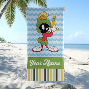 May include: A rectangular beach towel featuring a cartoon character with a green helmet and red shirt holding a yellow ray gun. The background has light blue waves, a green horizontal stripe with the text Your Name, and vertical stripes.