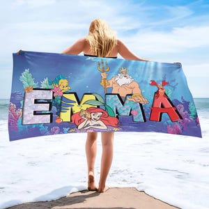 K&ouml;nnte beinhalten: Ein rechteckiges Strandtuch mit einer blauen Unterwasserszene, Korallen und Seetang. Der Name EMMA ist in gro&szlig;en, bunten Blockbuchstaben aufgedruckt. Das Design zeigt die Zeichentrickfiguren Arielle, K&ouml;nig Triton, Fabius und Sebastian.