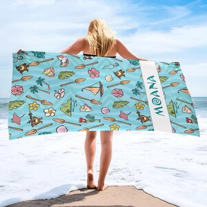 May include: A light blue beach towel with a repeating pattern of cartoon illustrations, including flowers, turtles, and boats. The word "MOANA" is printed vertically in white on a white stripe. The towel is held up by a person on a beach.