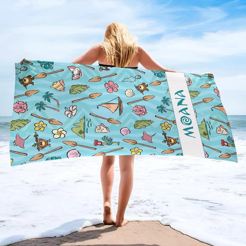 May include: A light blue beach towel with a repeating pattern of cartoon illustrations, including flowers, turtles, and boats. The word "MOANA" is printed vertically in white on a white stripe. The towel is held up by a person on a beach.