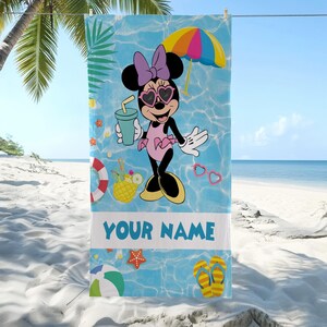 Op de afbeelding: Een strandlaken met Minnie Mouse in een roze badpak, hartvormige zonnebril en paarse strik. De blauwe achtergrond toont strandthema's, waaronder een parasol, drankje en de tekst "YOUR NAME".