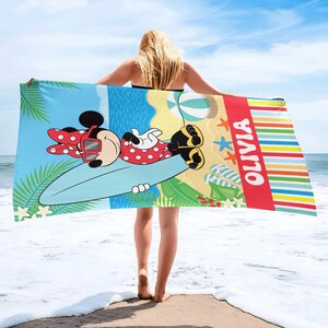 Puede incluir: Toalla de playa con Minnie Mouse con un vestido de lunares rojos y gafas de sol, tumbada en una tabla de surf. La toalla tiene una escena de playa colorida, el nombre "Olivia" y un fondo de cielo azul.