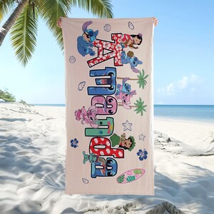 K&ouml;nnte beinhalten: Ein beiges Strandtuch mit dem Namen "Amara" in gro&szlig;en, bunten Buchstaben und Zeichentrickfiguren. Das Handtuch zeigt Bilder von Stitch, Lilo und tropischen Elementen wie Palmen und einem Surfbrett. Der Hintergrund zeigt eine Strandszene.