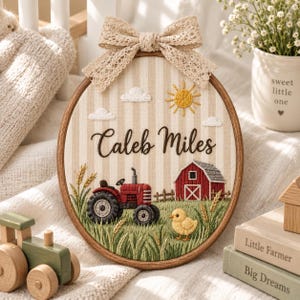Puede incluir: Un bastidor de bordado ovalado para pared con fondo de tela a rayas beige y blancas. El diseño incluye un tractor rojo, un granero rojo, un pollito amarillo, tallos de trigo, nubes y un sol amarillo. El nombre Caleb Miles está bordado en negro.