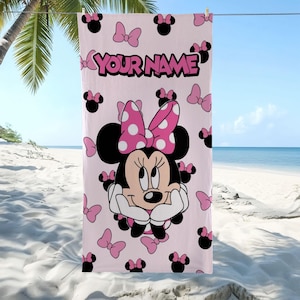 May include: A light pink beach towel featuring a large graphic of Minnie Mouse with a polka-dot bow, surrounded by smaller black mouse ear silhouettes and pink bows. The text "YOUR NAME" is printed in bold pink letters at the top center.