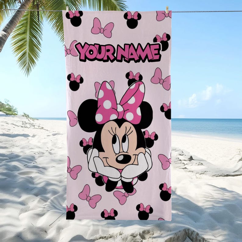 May include: A light pink beach towel featuring a large graphic of Minnie Mouse with a polka-dot bow, surrounded by smaller black mouse ear silhouettes and pink bows. The text "YOUR NAME" is printed in bold pink letters at the top center.