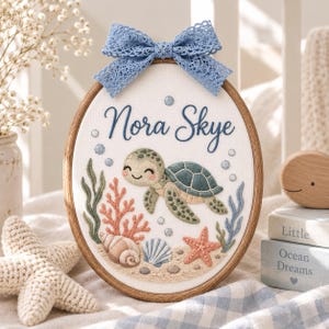 以下が含まれることがあります： 白い布地に青い筆記体でNora Skyeと刺繍された楕円形の刺繍枠。緑色のウミガメ、サンゴ、貝殻、ヒトデが刺繍されており、上部には青いレースのリボンがあしらわれています。